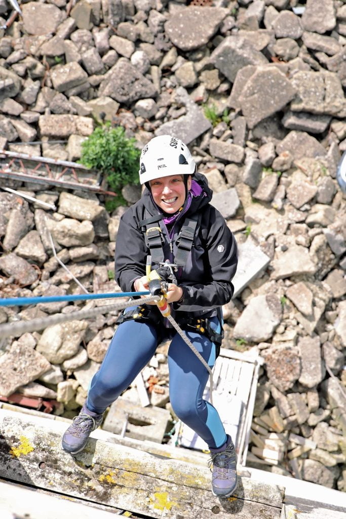 Smiling woman in a white safety helmet holding blue and white safety ropes while abseiling down a tower block